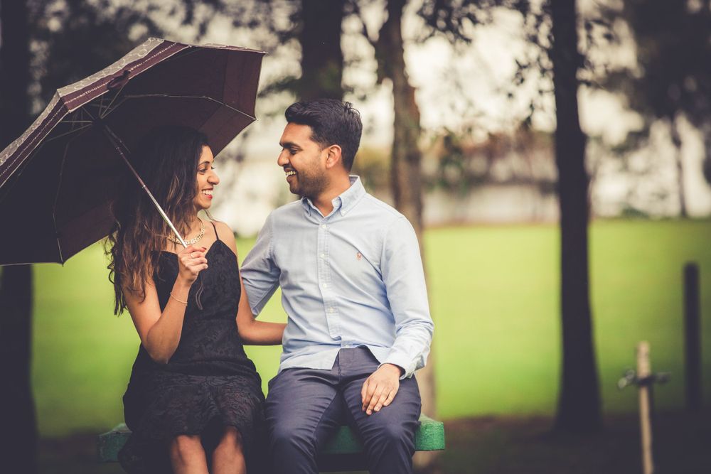 Pre Wedding shoot of a Sri Lankan Tamil couple sitting facing each other in the rain with a black umbrealla in Sydney