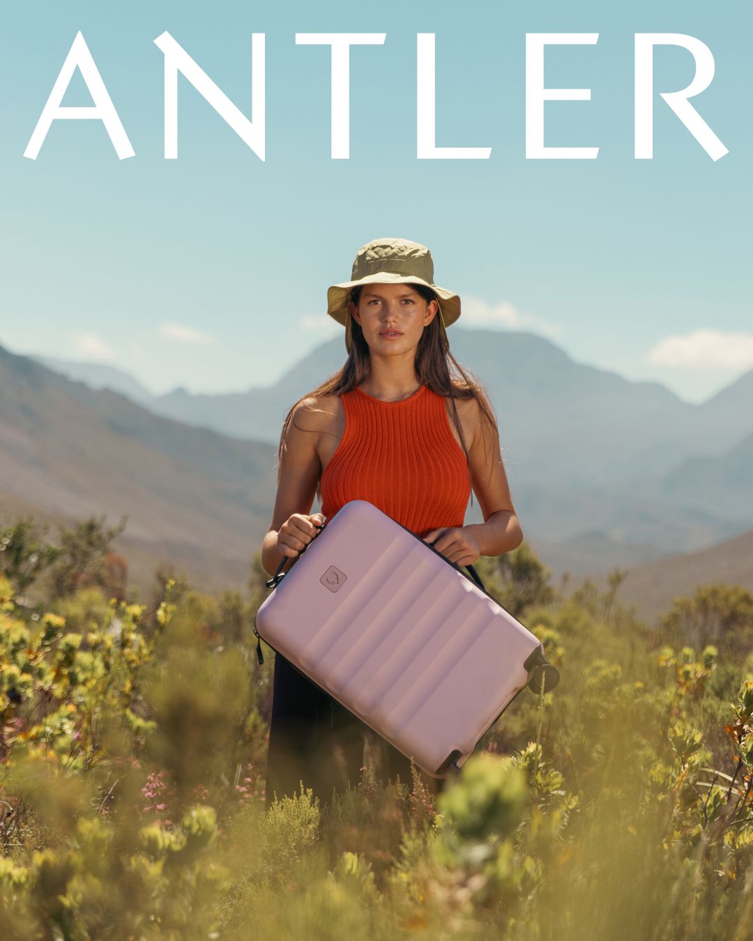 Woman in red top holding purple Antler suitcase in mountainous desert landscape.