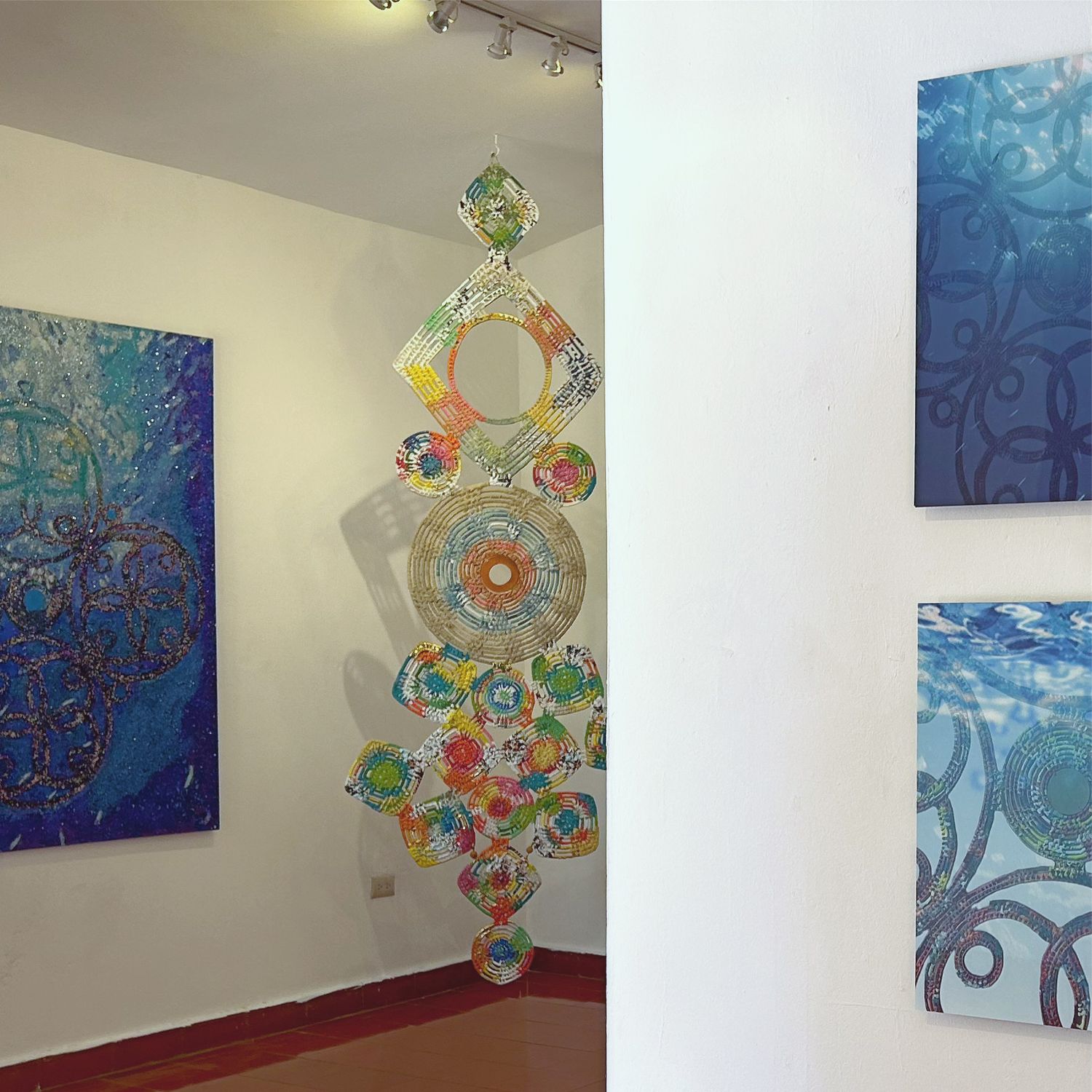 Modern gallery space showing blue paintings and suspended circular sculpture with colorful details.