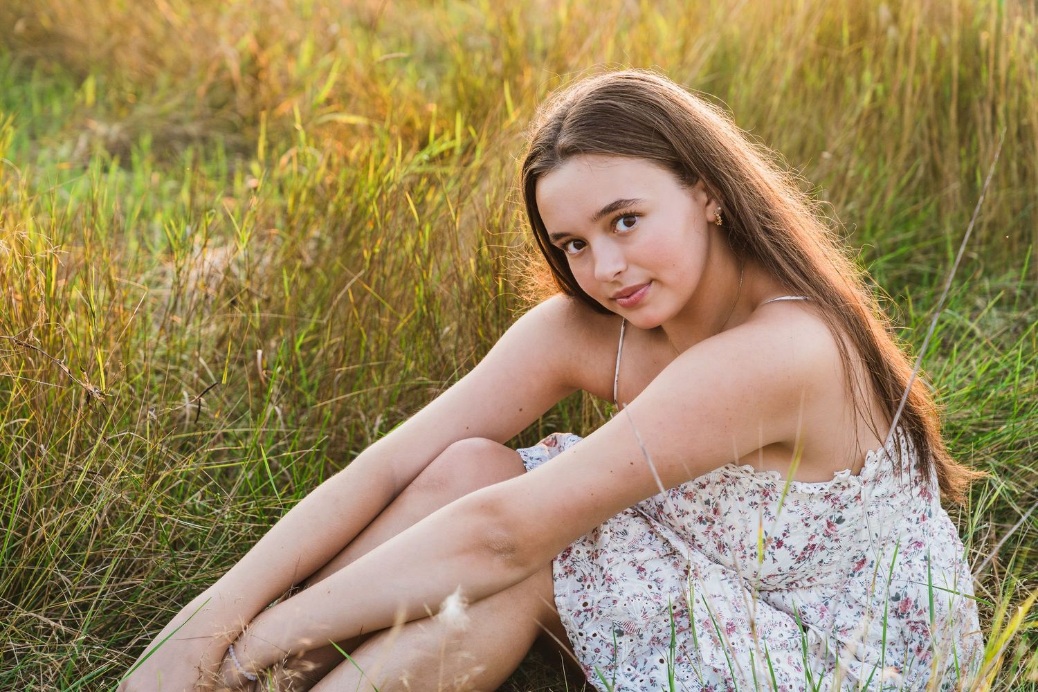 A person in a floral dress sits in tall grass during golden hour sunset lighting.