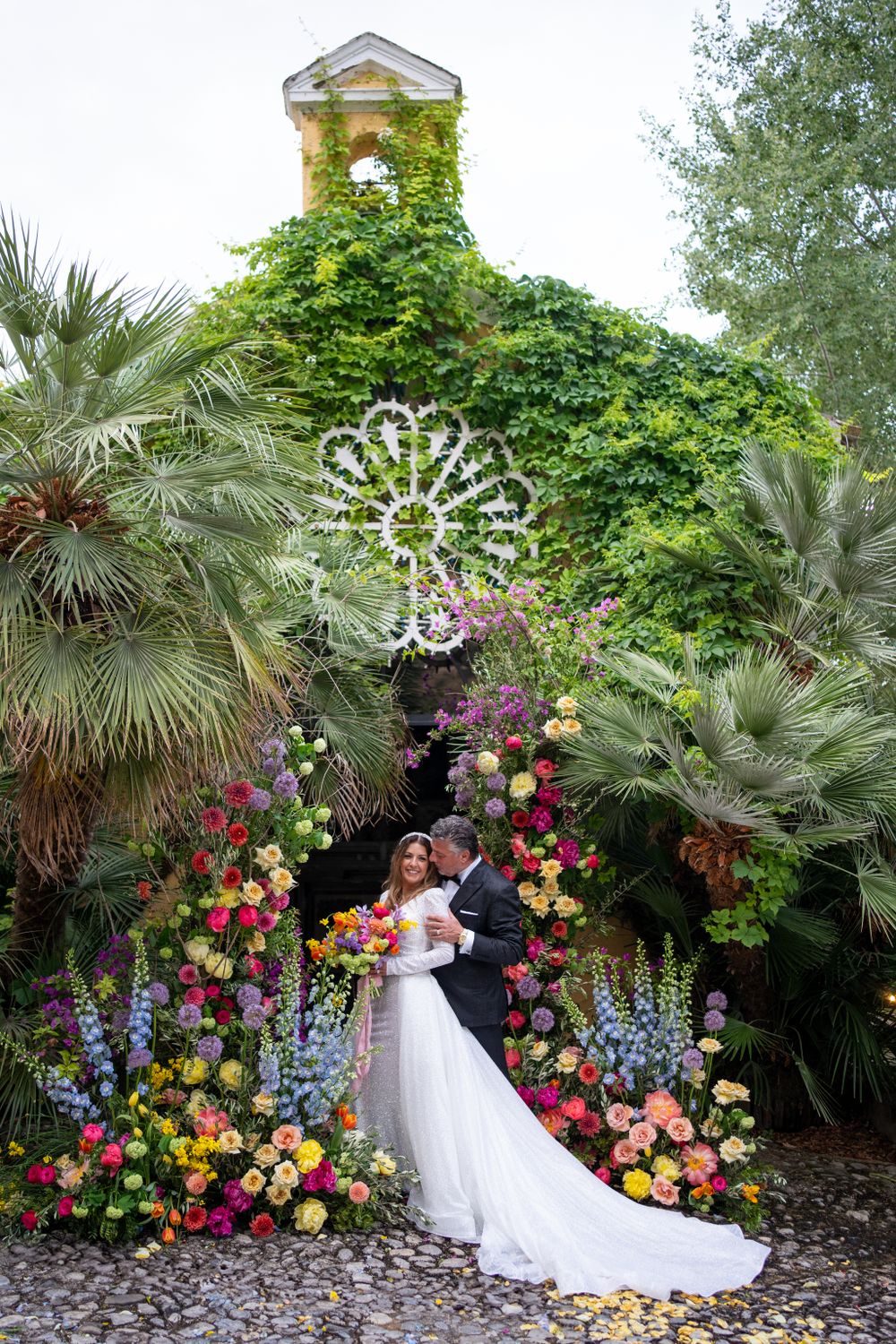 Lush garden wedding scene with colorful floral arch and tropical palm trees.