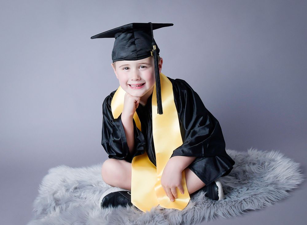 Nursery graduation image, in cap and gown sitting on a grey furry carpet. Studio light with a grey backdrop.