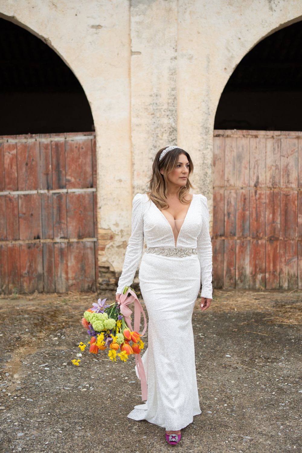 Bride in white dress holds colorful bouquet while posing against rustic wooden doors.