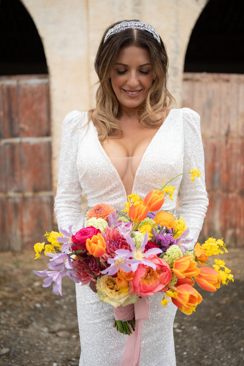 Bride holds vibrant spring bouquet with orange, yellow and purple flowers against stone backdrop.