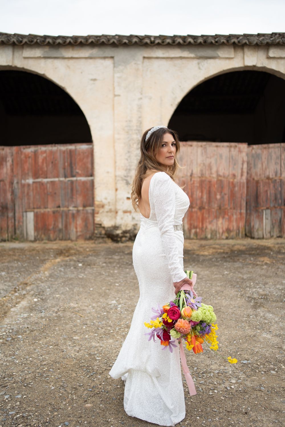 Bride in white dress poses with colorful bouquet against rustic architectural background.