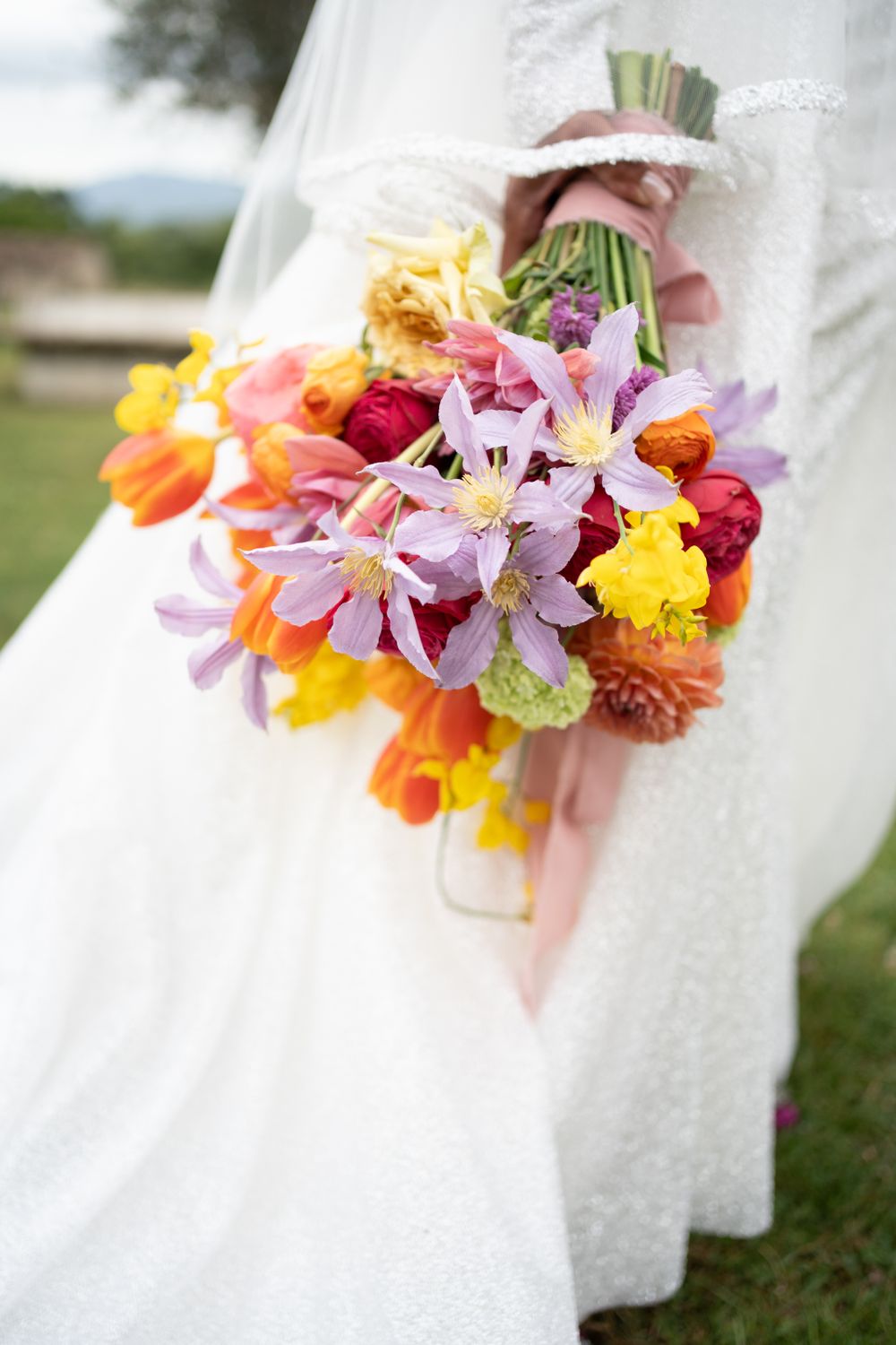Vibrant wedding bouquet with pink, orange, and yellow flowers against a white dress background.