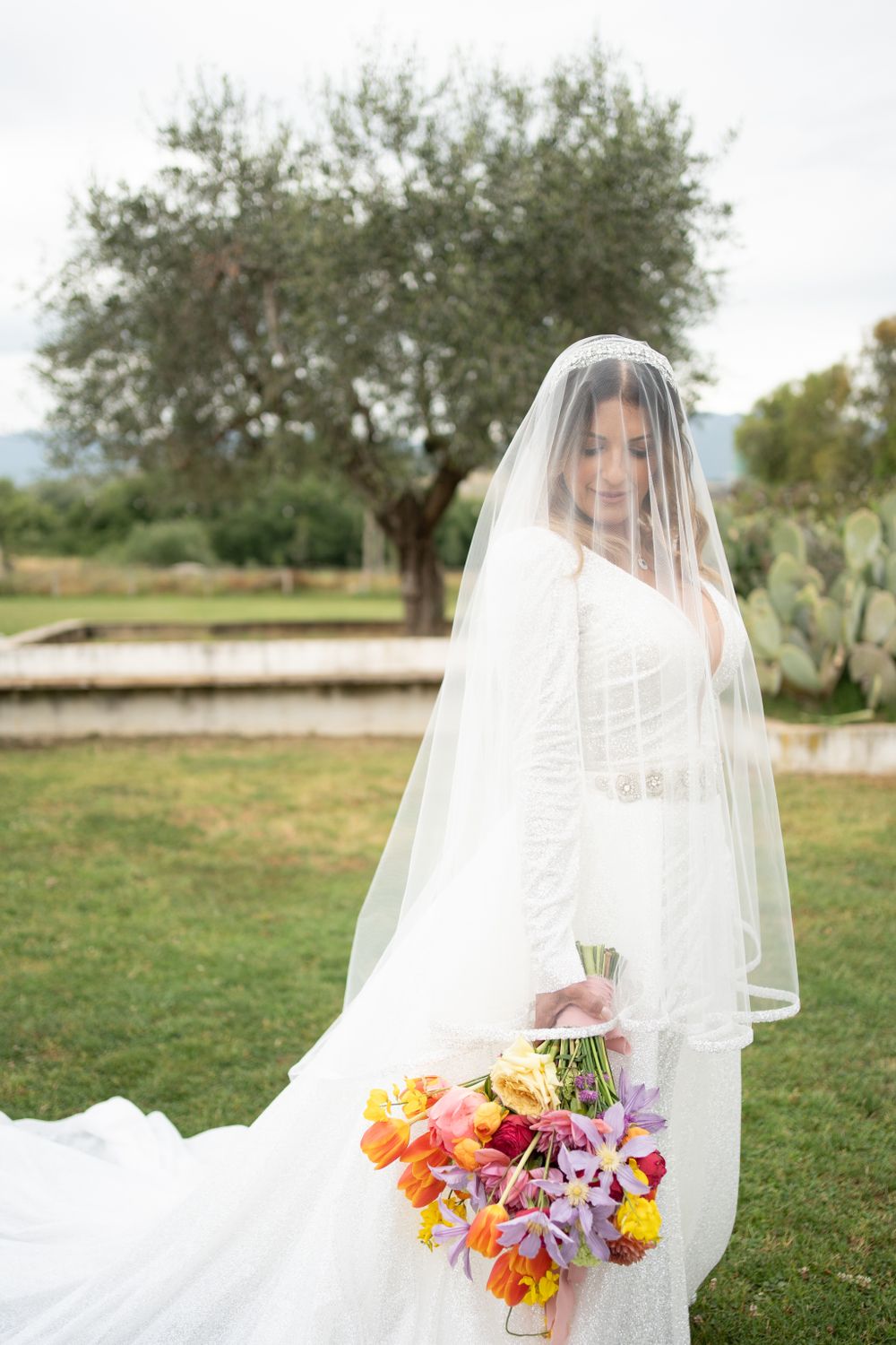 Ethereal bridal portrait with flowing white veil and colorful bouquet in an outdoor garden setting.