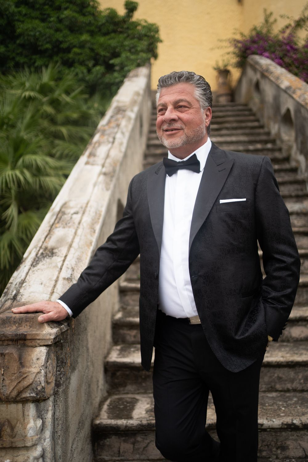 Groom in classic black tuxedo poses on stone steps surrounded by lush greenery.