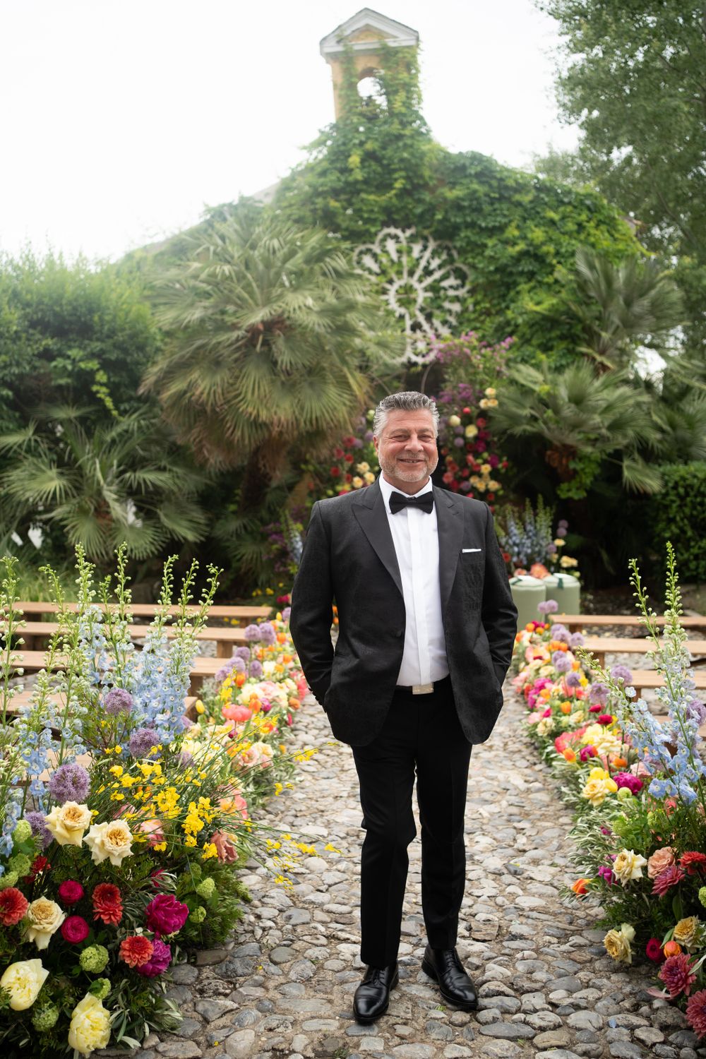 Groom in black tuxedo stands among colorful garden flowers and palm trees.