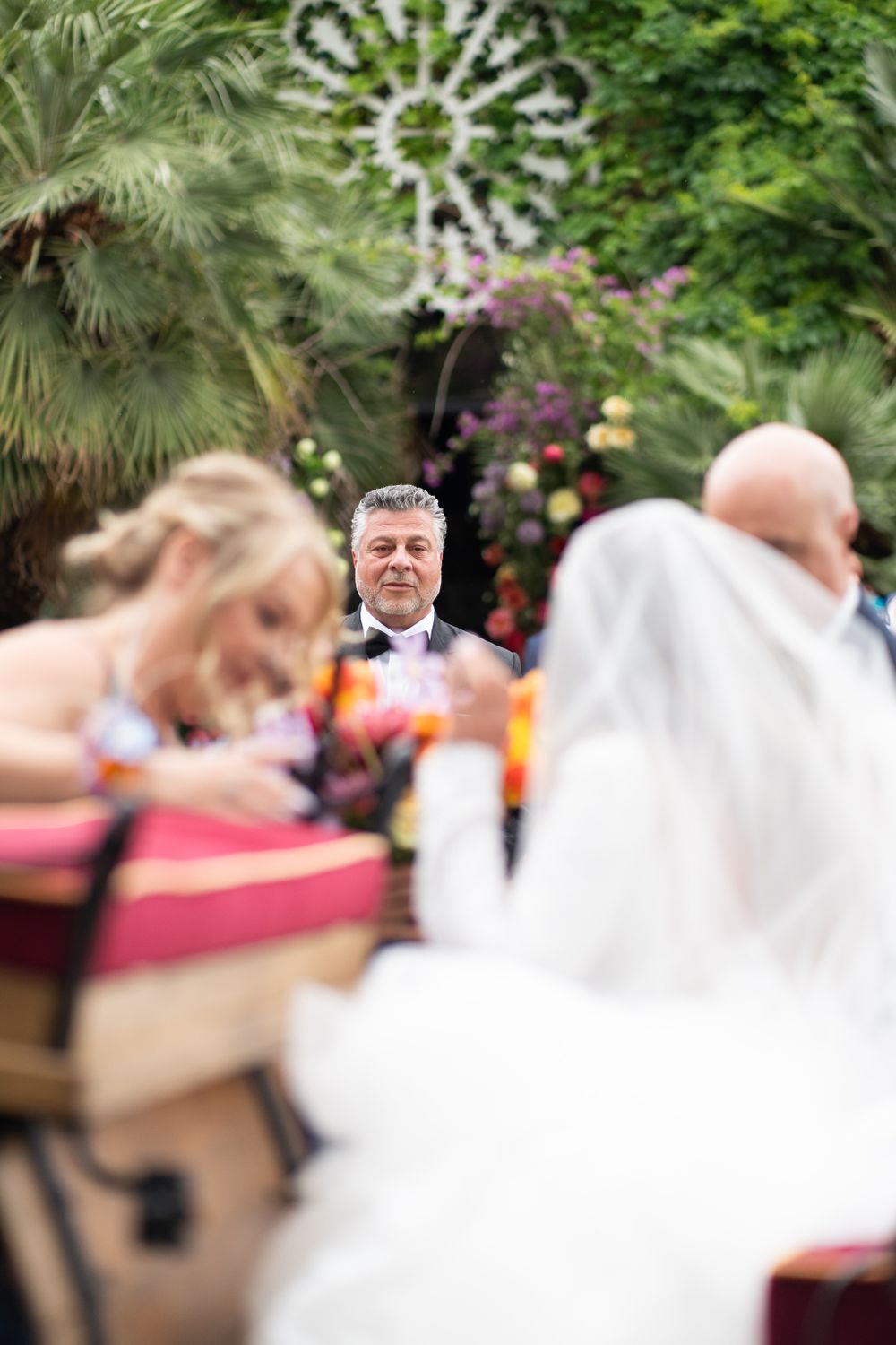 Wedding ceremony scene with blurred figures and tropical greenery in the background.