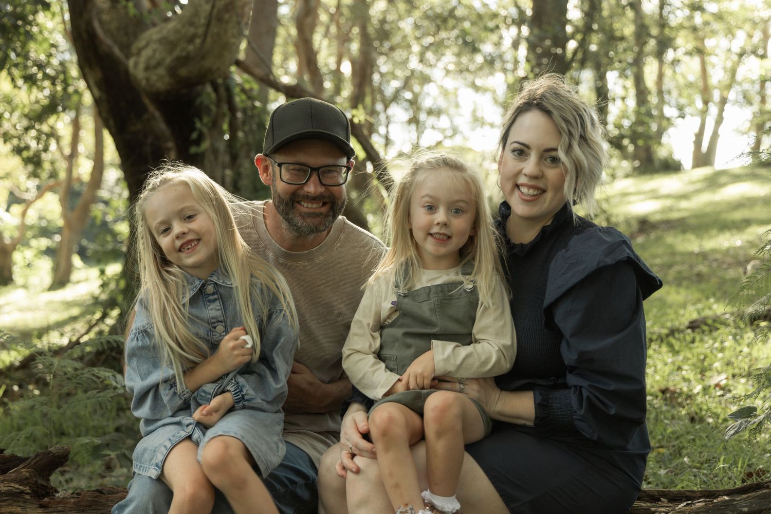 A happy family of four sits together on rocks in a wooded outdoor setting.