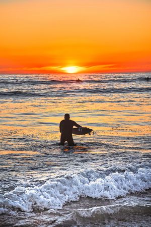 A silhouetted person holds a surfboard in the ocean while watching a vibrant orange sunset over crashing waves.