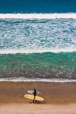 A surfer carrying a yellow longboard walks along a sandy beach with turquoise waves crashing in the background.