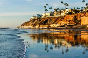 Luxury homes perched on cliffs above a serene beach with palm trees reflected in calm ocean waters during golden hour.