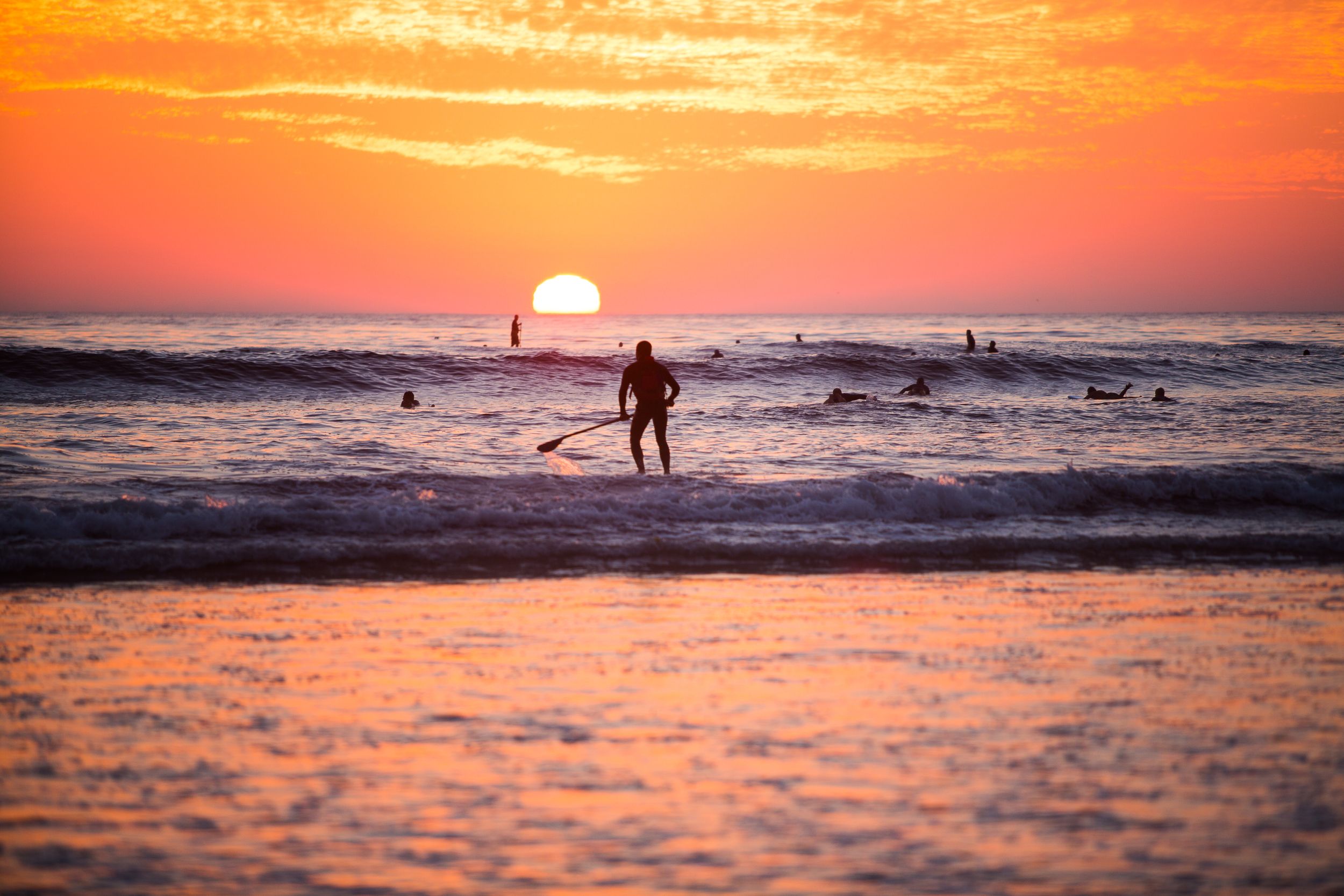 Silhouette of surfer at beach during dramatic orange sunset over ocean waves.