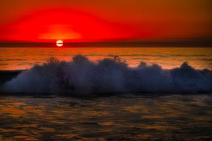 Ocean waves crash dramatically against a vibrant red sunset sky creating a stunning silhouette effect over the water.