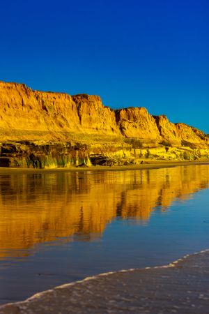 Dramatic yellow coastal cliffs reflecting in calm ocean waters against deep blue sky.