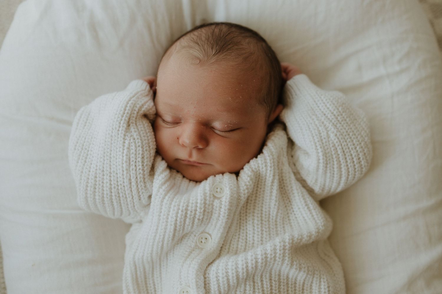 A sleeping baby wearing a white knit sweater rests peacefully on white bedding.