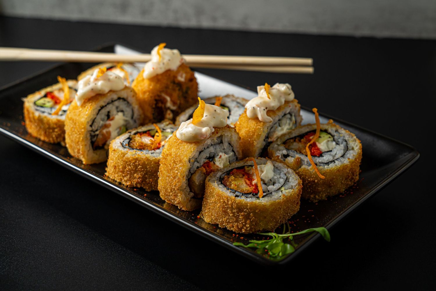 Crispy tempura sushi rolls garnished with sauce and tempura flakes served on a dark plate.