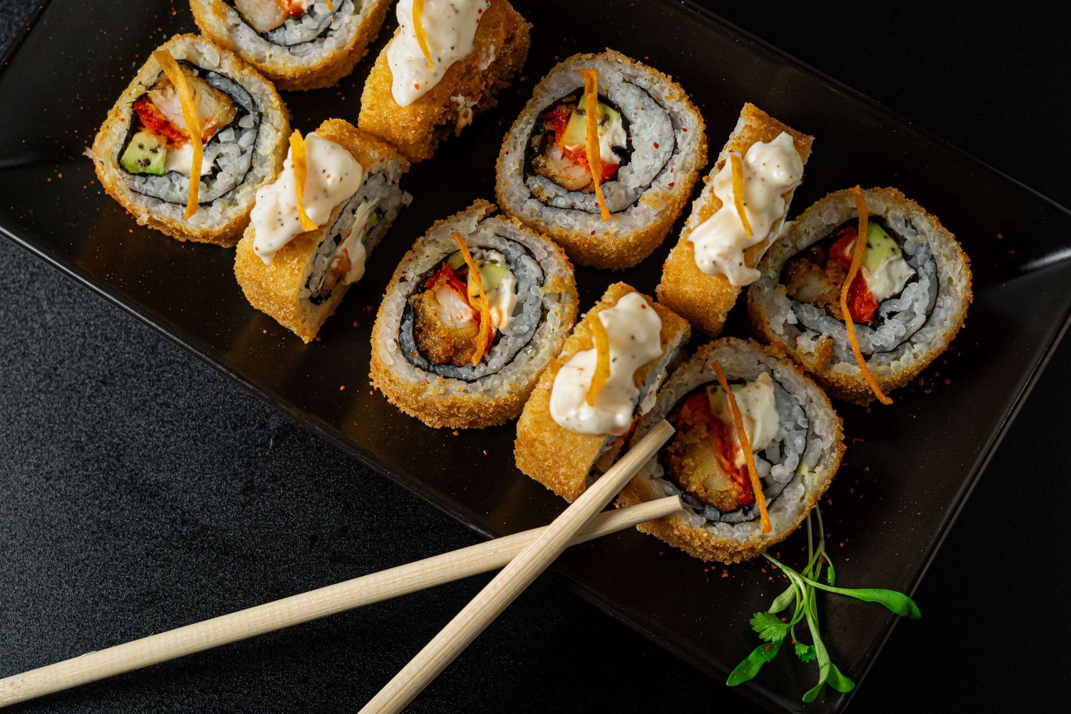 Delicious tempura sushi rolls arranged on black plate with chopsticks and garnish.