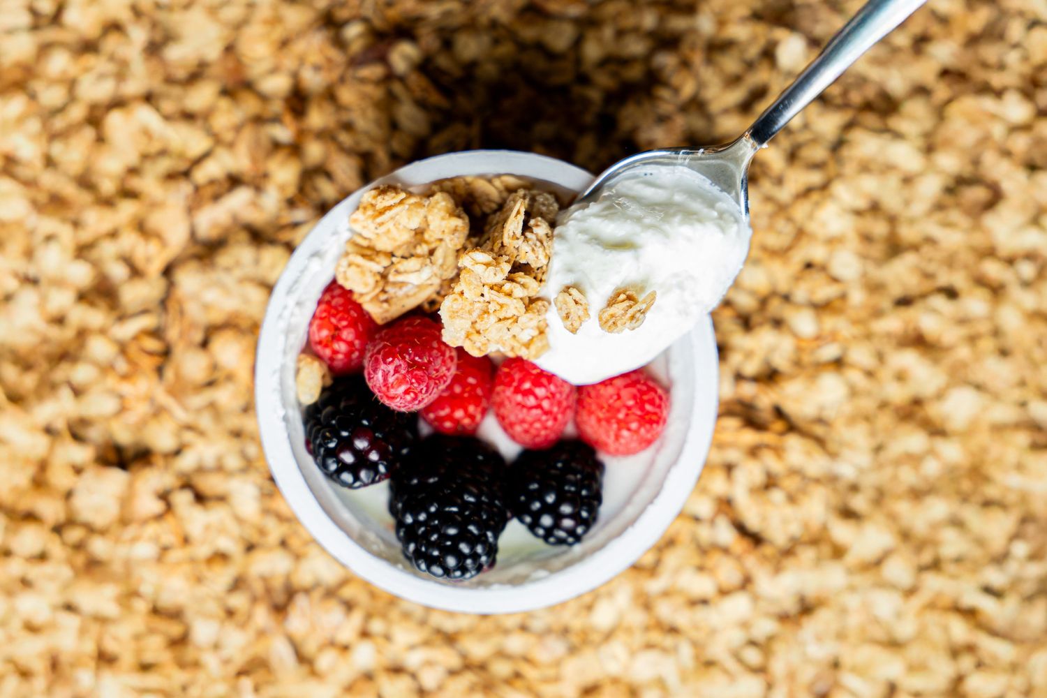 A yogurt parfait topped with granola, fresh raspberries and blackberries viewed from above with a spoon.