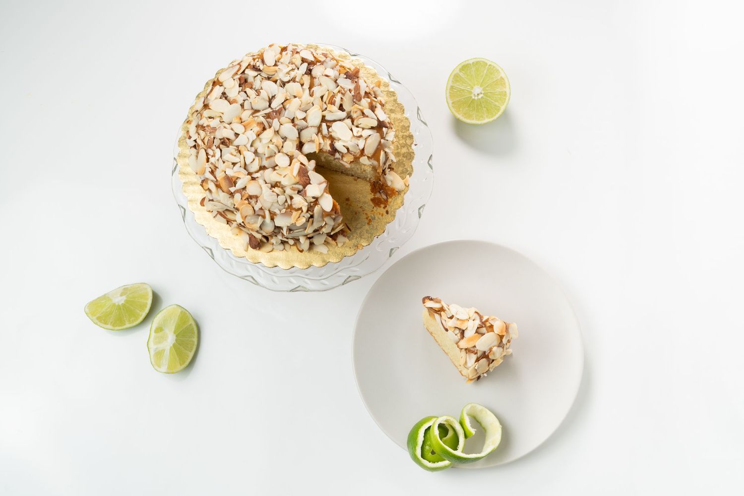 Almond-topped cake on cake stand with lime wedges on white background.
