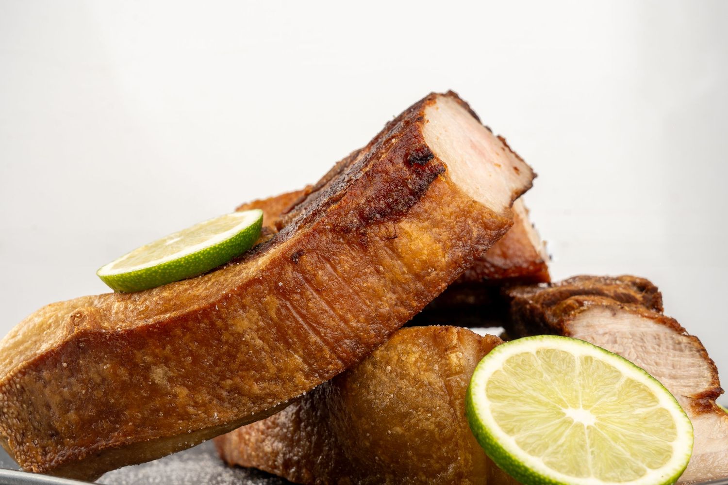 Thick slices of crusty chicharron garnished with lime wedges on light background.