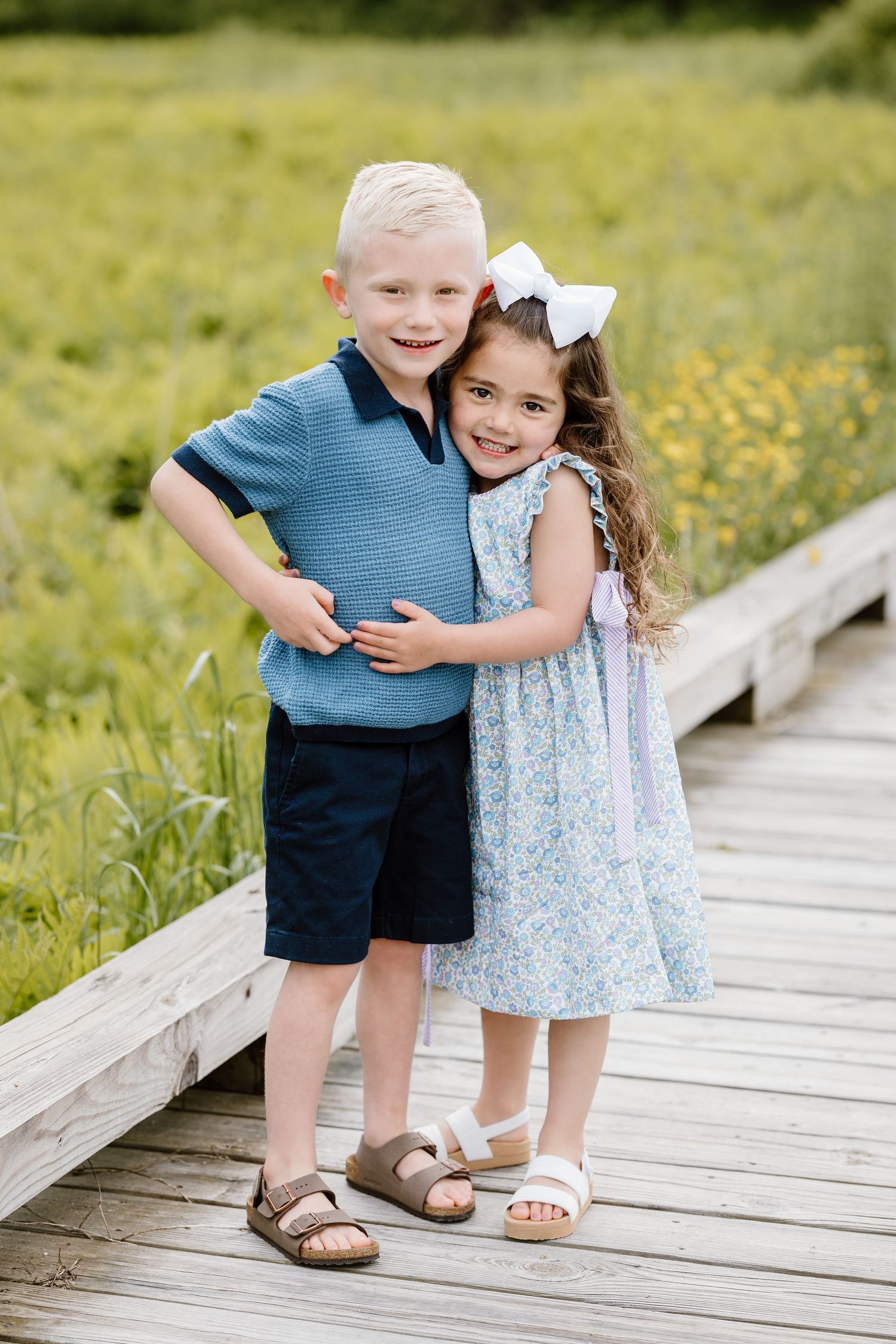 Two young children in summer outfits standing together and smiling in a sunlit field.