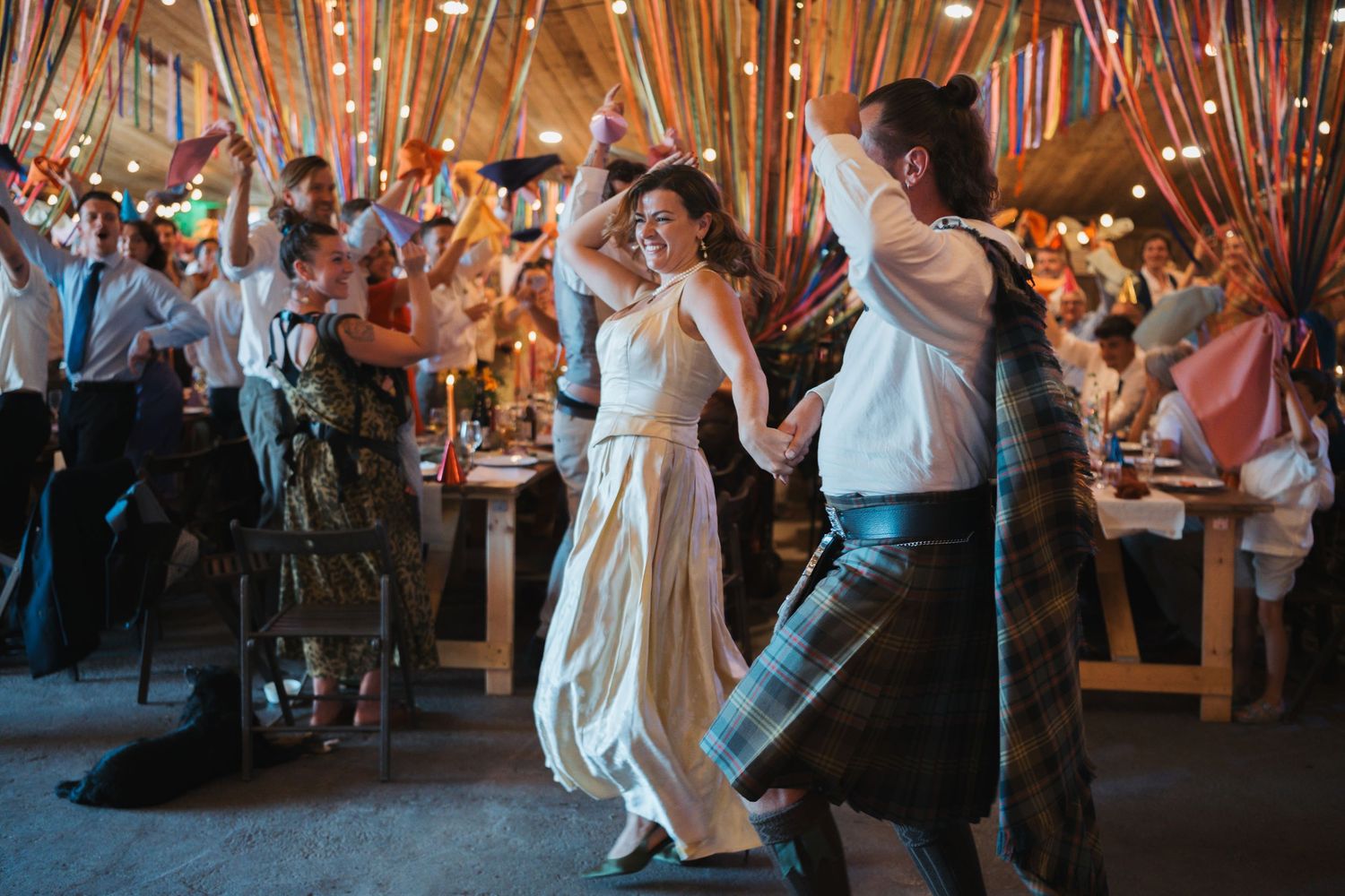 Wedding guests dancing energetically on white dance floor under draped ceiling with lights.