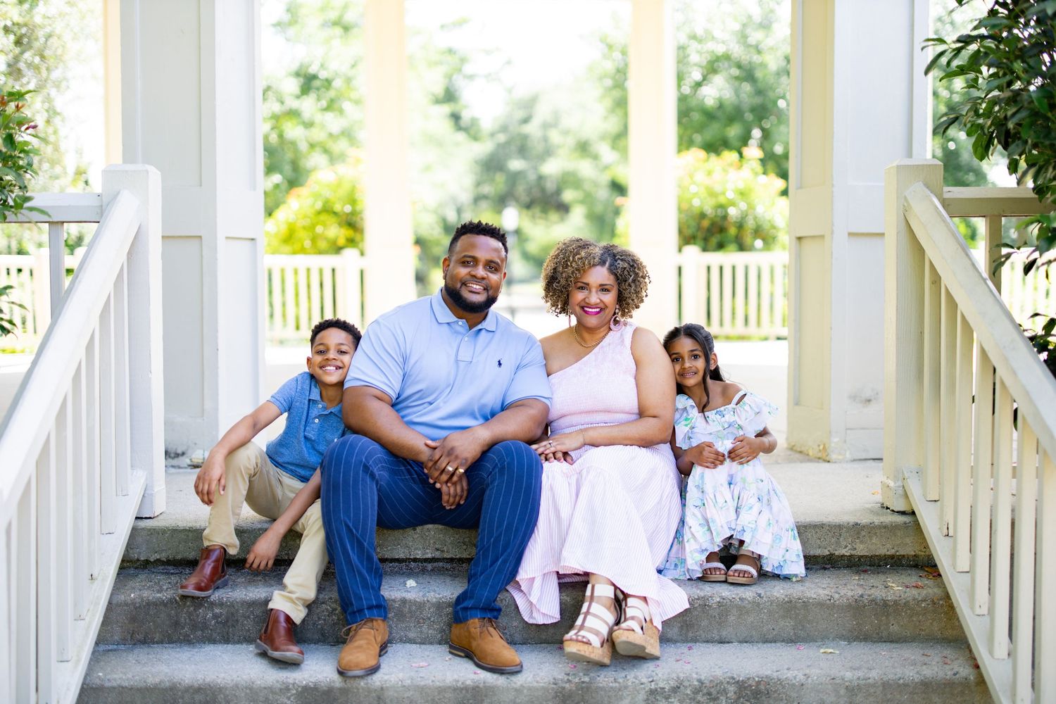 A loving family sits together on white porch steps during a sunny outdoor photo session.