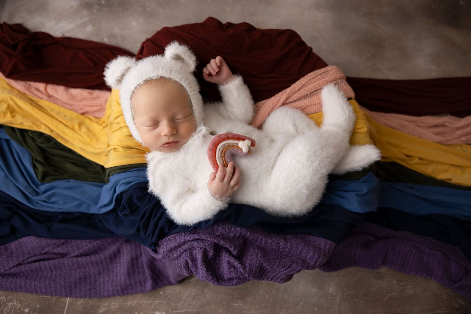 Sleeping baby wearing white fuzzy outfit on colorful striped fabric background.