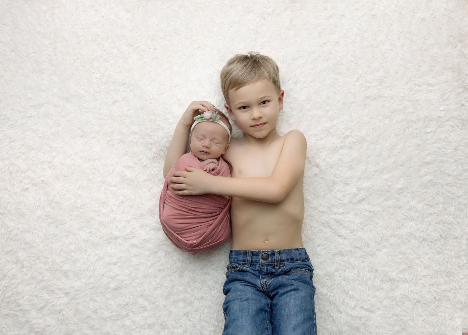 Sweet sibling portrait with baby in pink wrap lying on white backdrop.