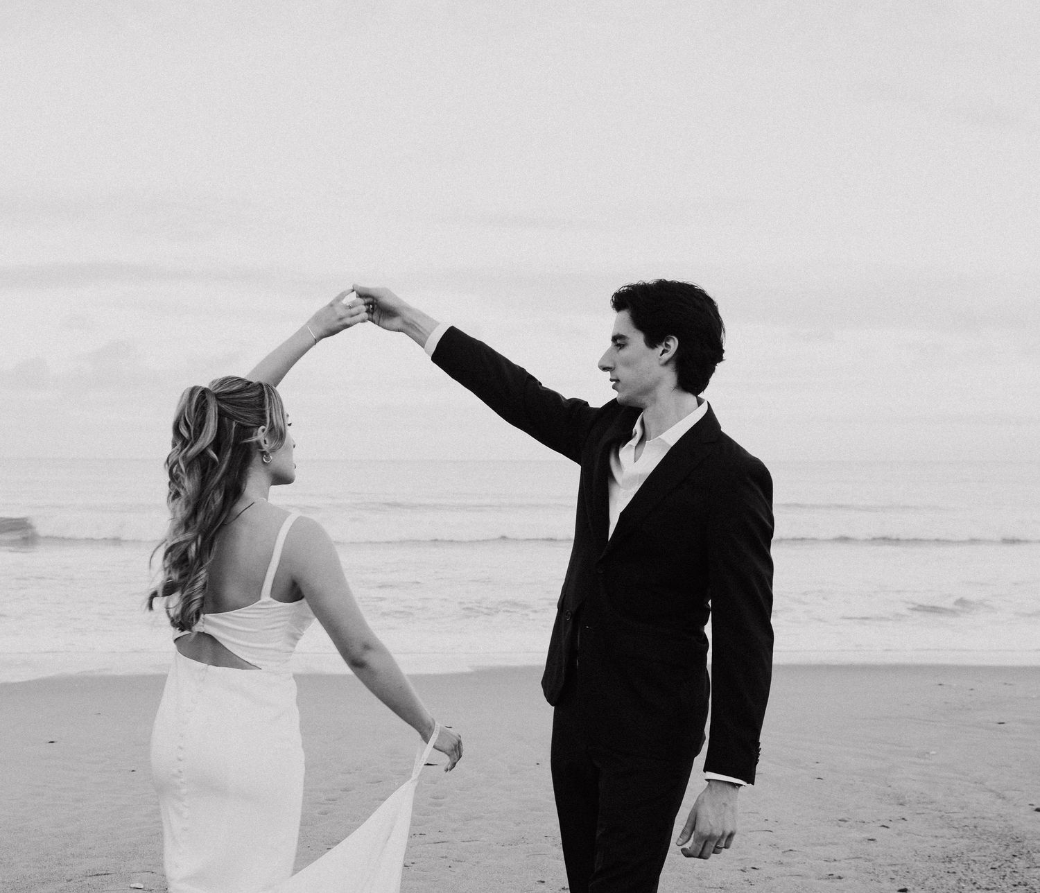 A couple in wedding attire dances playfully together on a beach at sunset.