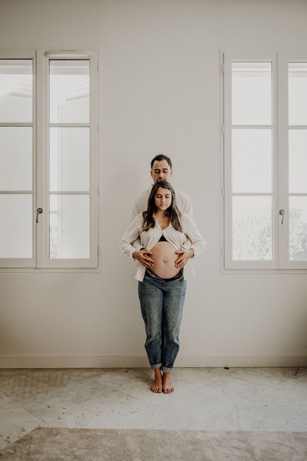 Maternity photo of an expectant couple in a bright white room with large windows.photographe futur maman Bordeaux, Gironde