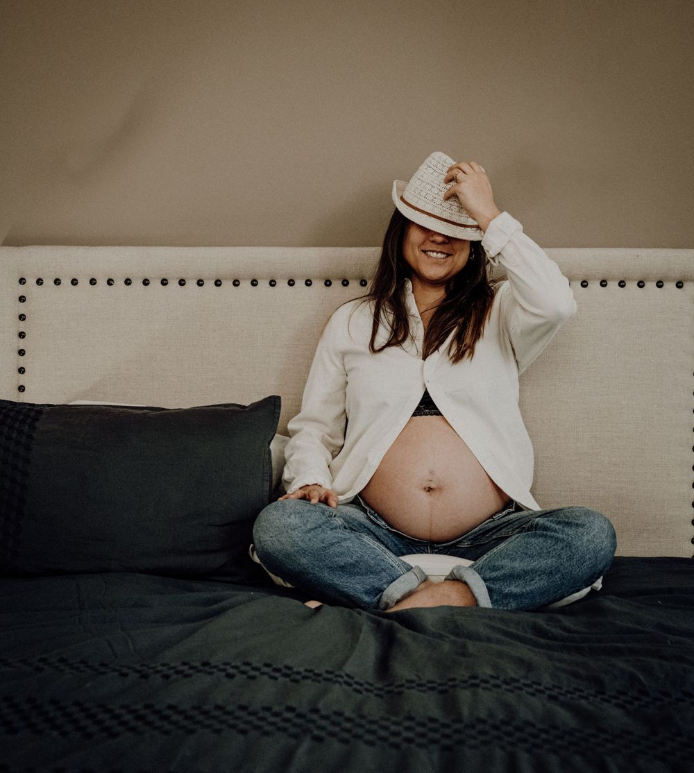 Pregnant woman in jeans and white shirt sitting cross-legged on a beige headboard bed. photographe grossesse bordeaux