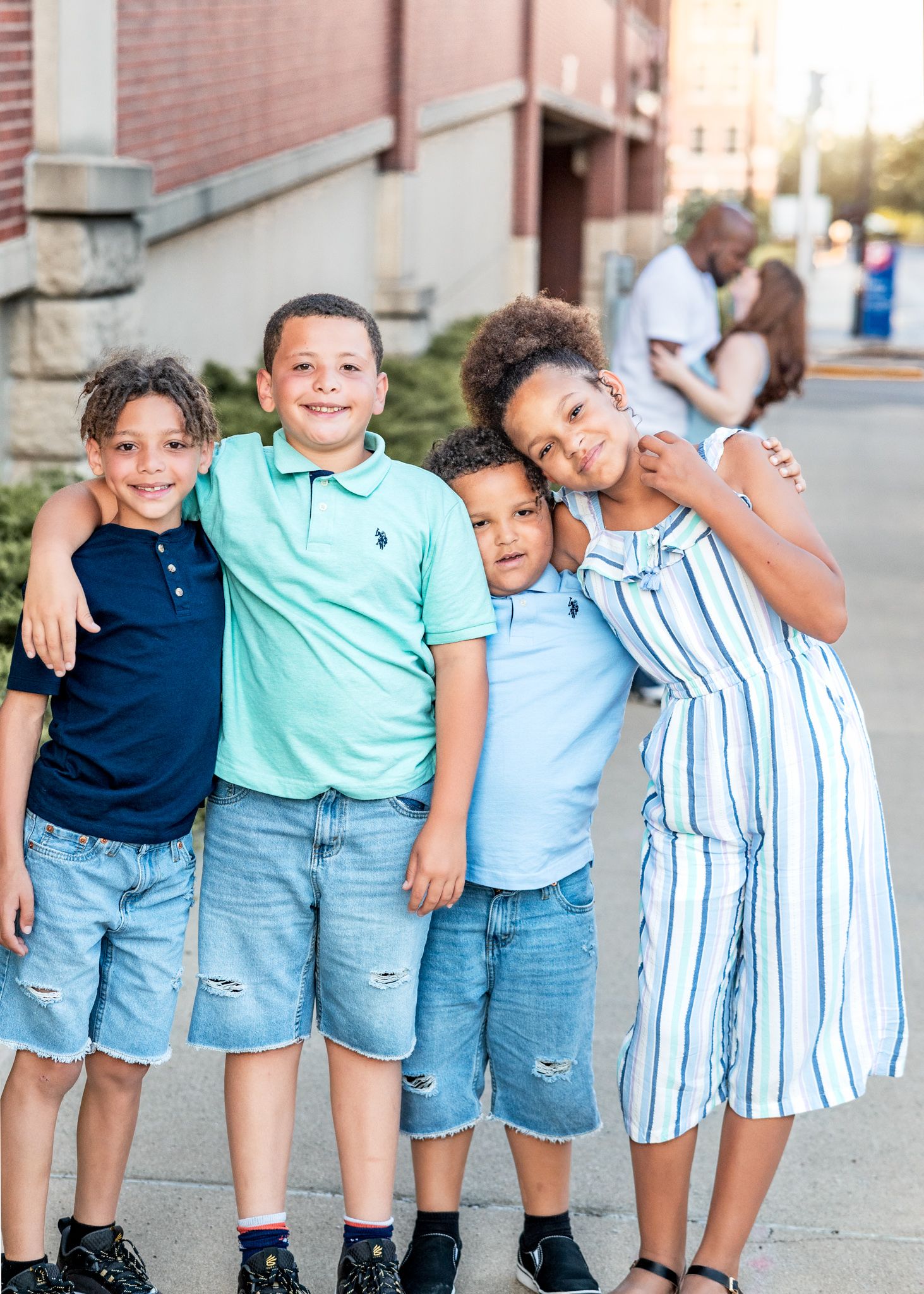 Four children stand together in casual summer clothing on a sidewalk outside a brick building.
