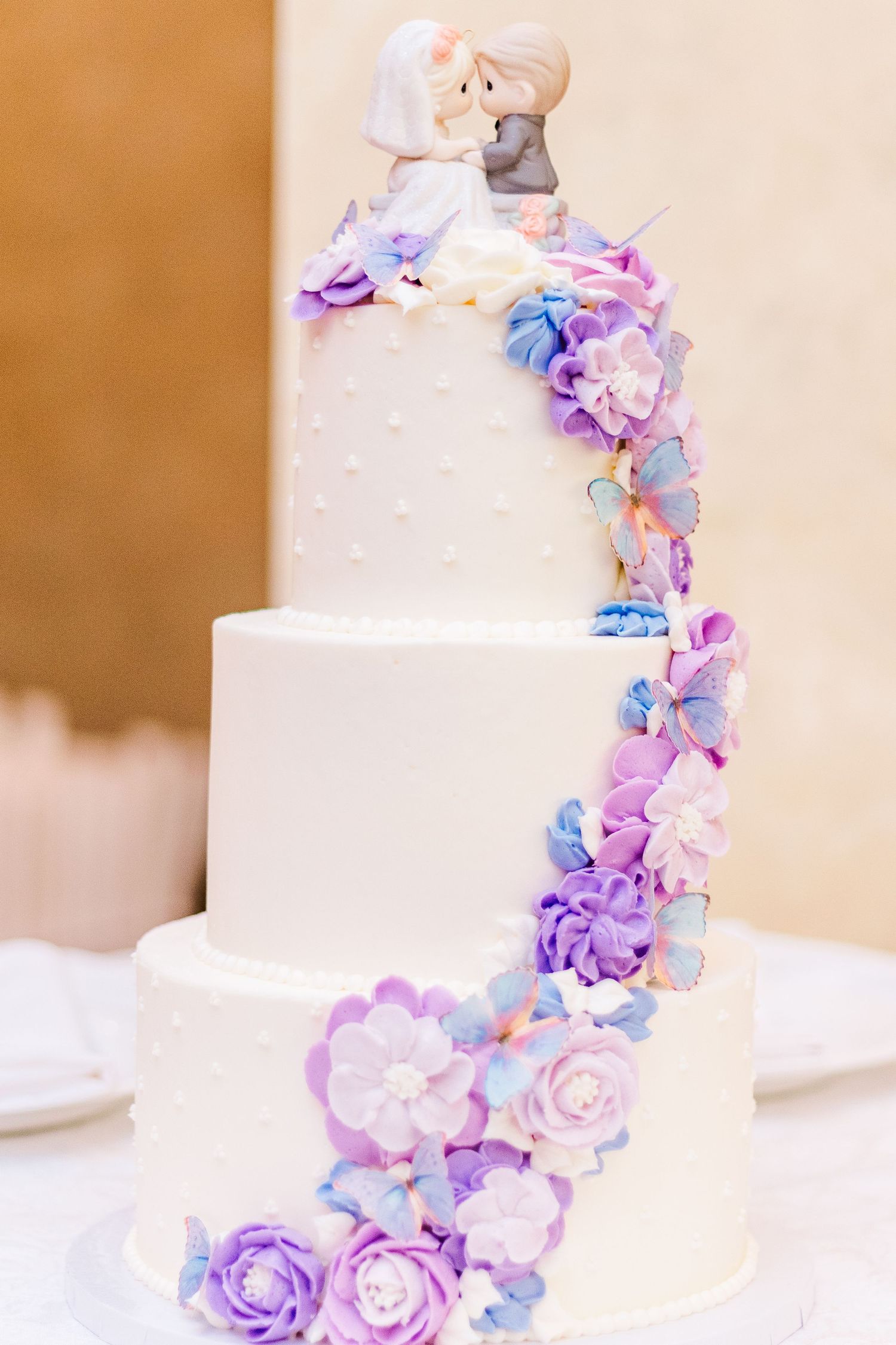 White wedding cake decorated with cascading purple and blue flowers and bride and groom topper.