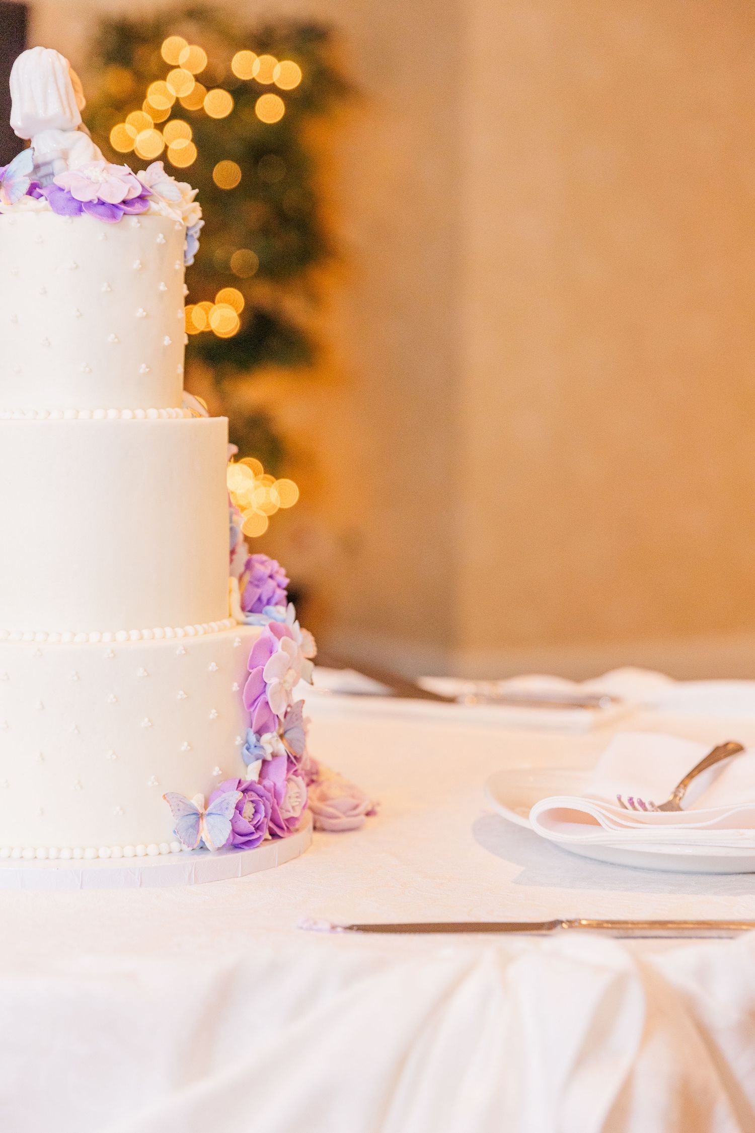 Elegant white tiered wedding cake with purple floral accents against warm bokeh background.