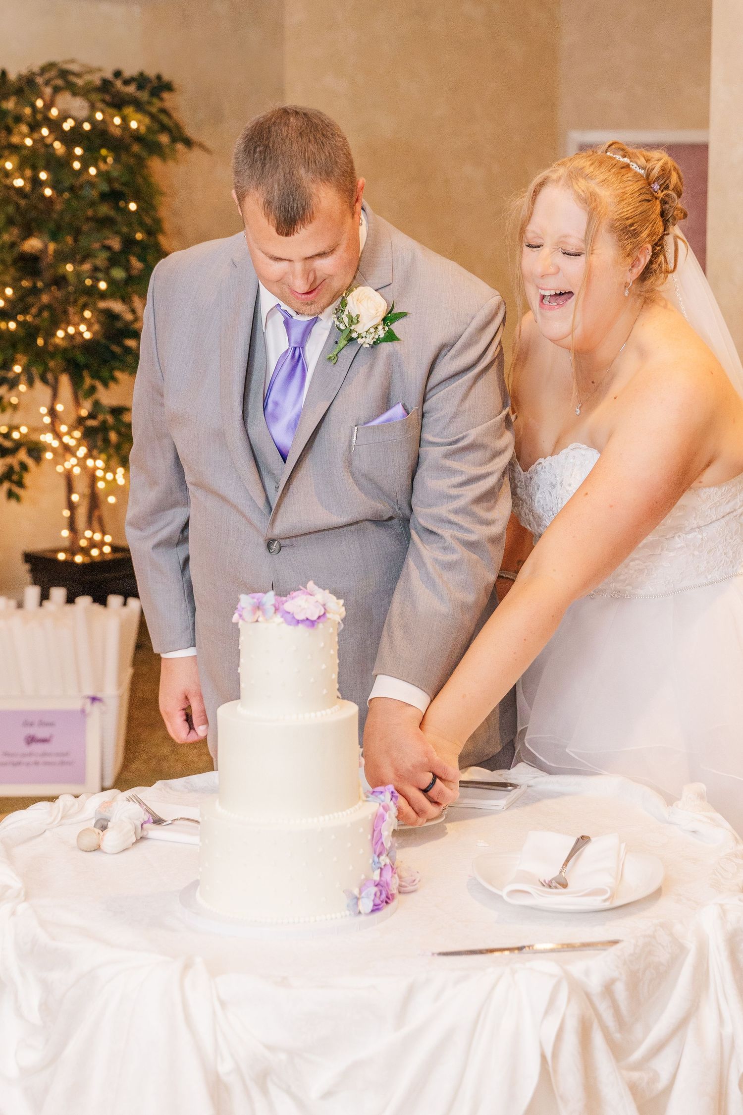 Couple cutting their white wedding cake decorated with purple flowers at reception.
