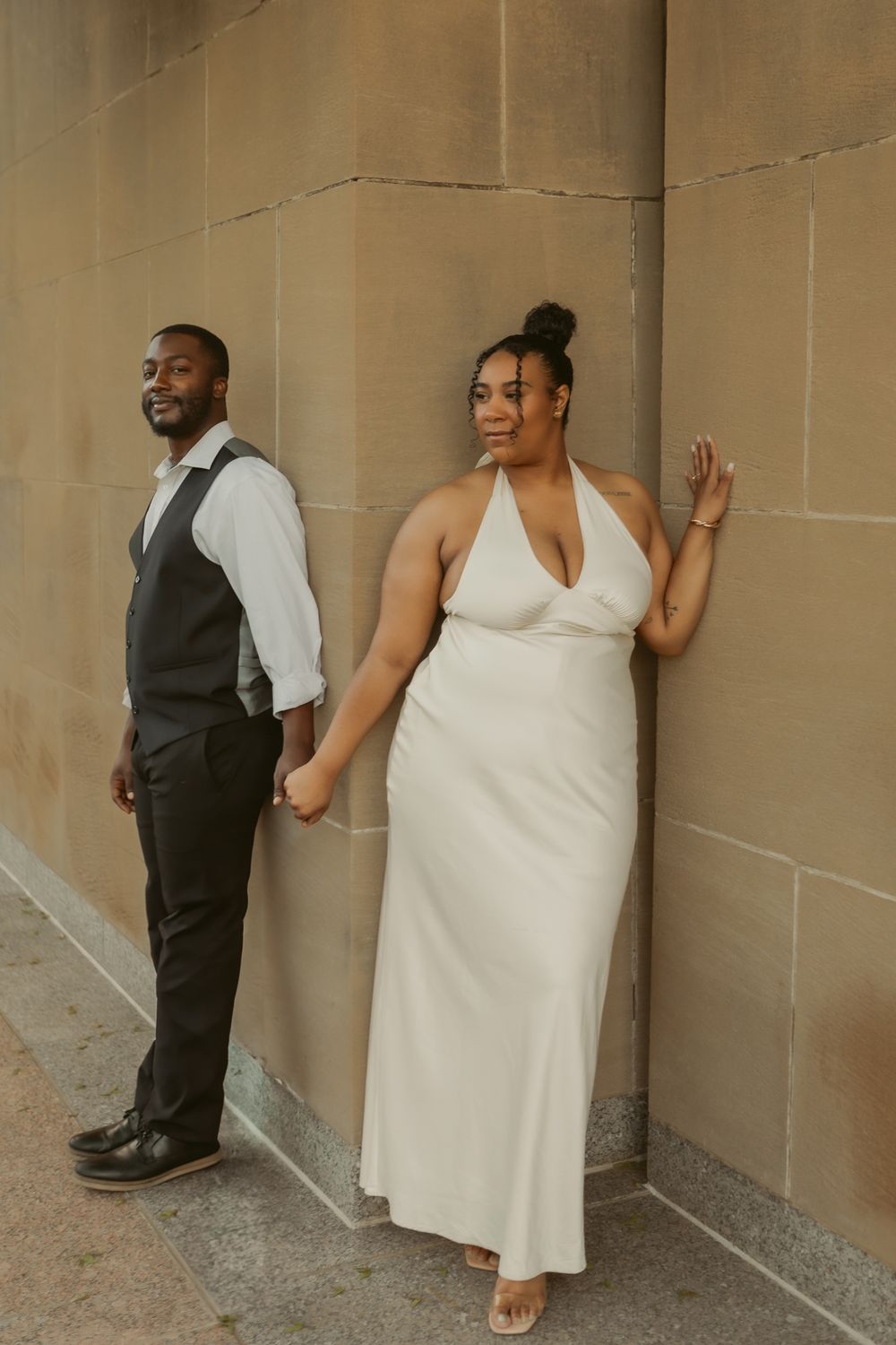 A couple in formal attire poses against a neutral stone wall, with him in a vest and her in a halter-style white dress.