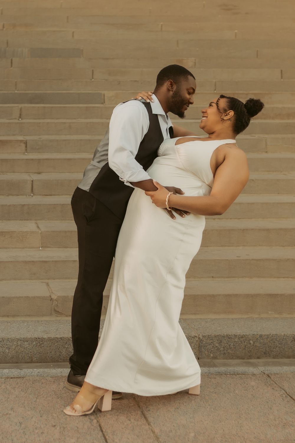 Wedding couple in formal attire share an intimate dance pose against a textured beige wall.