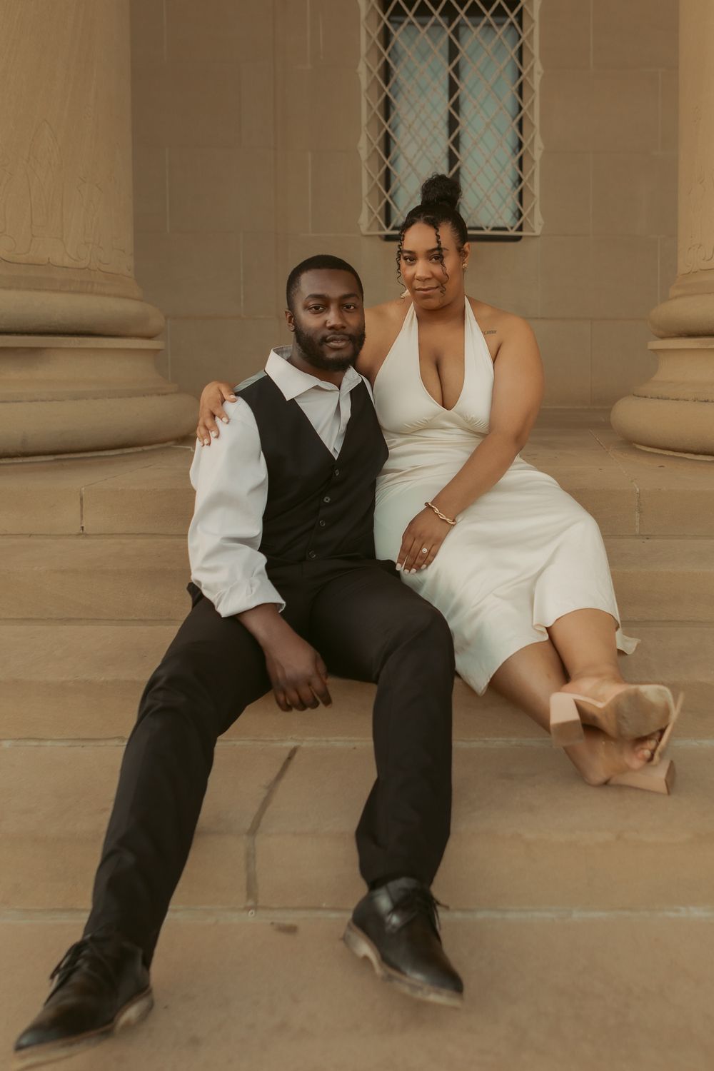 Wedding couple in coordinated black and white attire sitting together on stone steps by a classic building.
