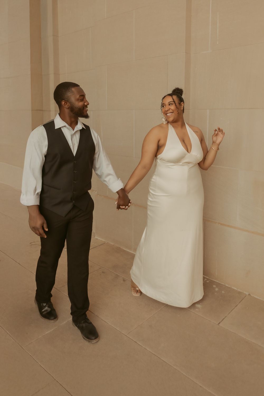 A couple holds hands while posing in formal attire against a beige wall, showing joy and laughter.