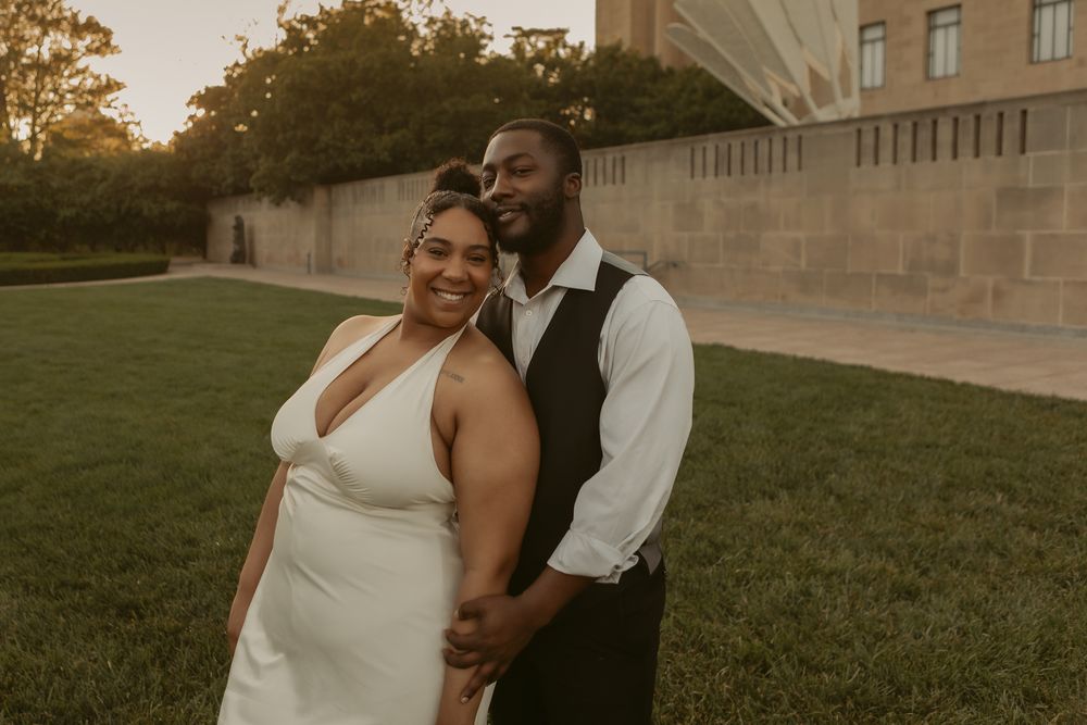 A happy couple in wedding attire poses together outdoors at sunset near a stone building with manicured lawns.
