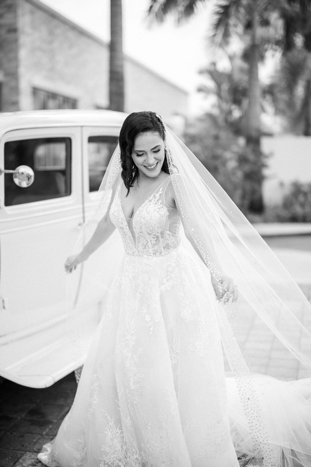 Black and white portrait of bride in flowing lace gown next to vintage car.