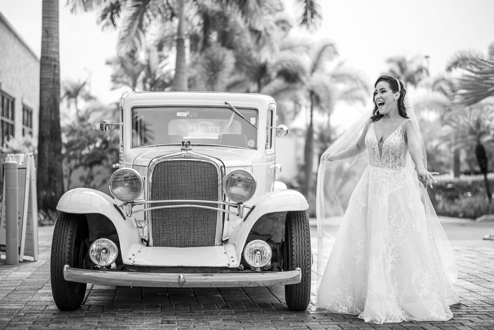 Bride in elegant lace gown poses beside classic white vintage car.