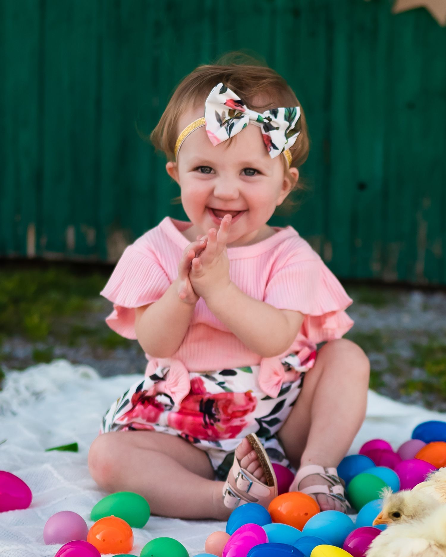 A toddler in pink with a floral headband sits among colorful plastic eggs.