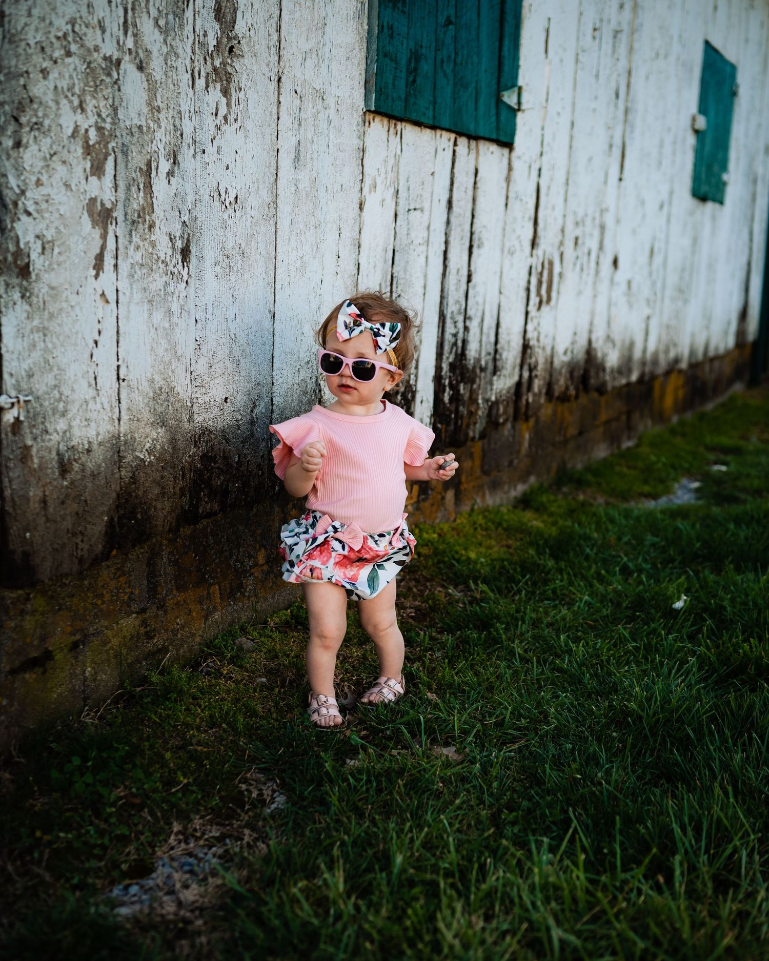 A toddler in pink wearing sunglasses dances by a rustic white wall.