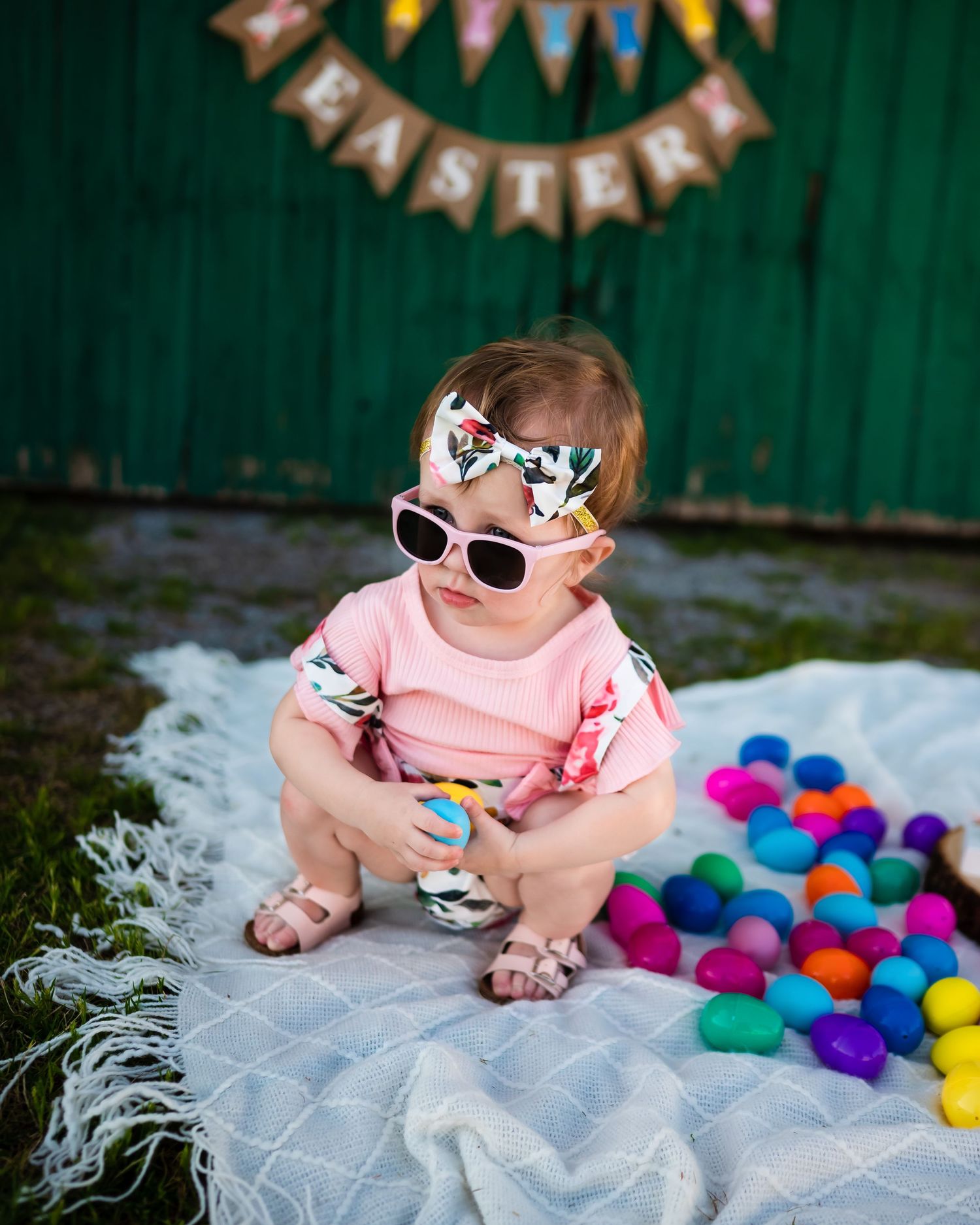 A toddler in pink wearing sunglasses sits among colorful Easter eggs.