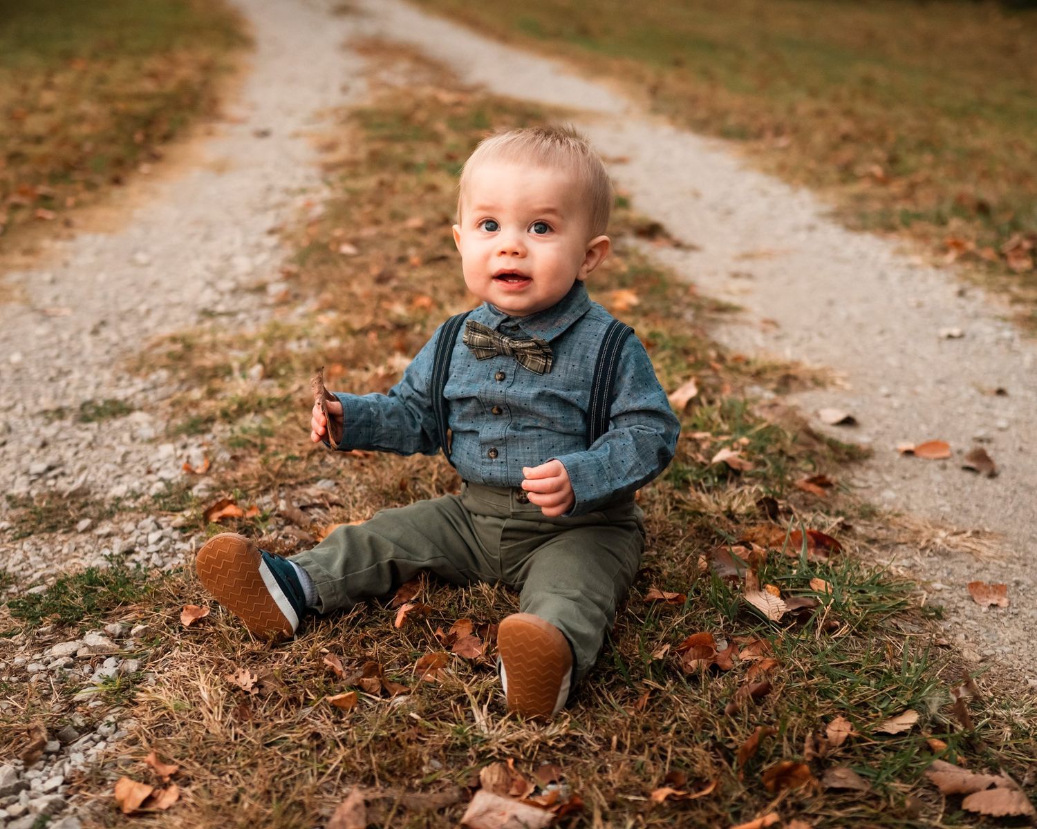 Baby in denim and green jacket sitting among autumn leaves on dirt path.